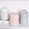 A set of three compact desktop trash cans in white, pink, and gray with flip covers.
