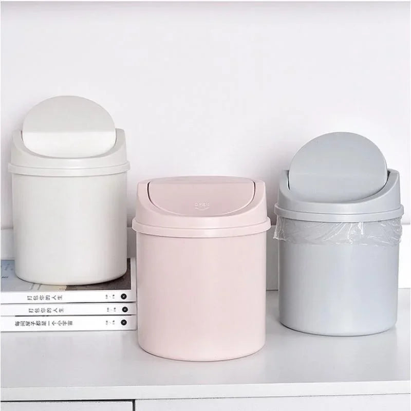 A set of three compact desktop trash cans in white, pink, and gray with flip covers.