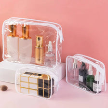 A set of three transparent plastic travel organizer bags containing gold and clear cosmetic bottles on a pink background.