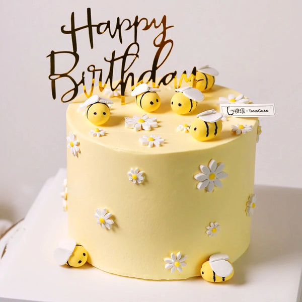A round yellow cake decorated with white daisy accents and small yellow bee toppers, topped with black script reading Happy B