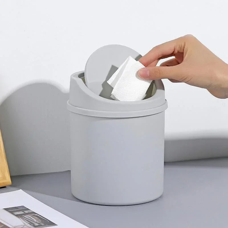 A hand is lifting a white paper towel from the flip cover of a gray compact desktop trash can.