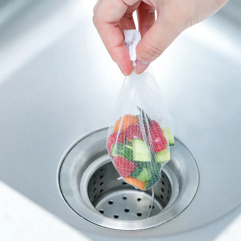 A hand is placing a clear disposable filter bag filled with colorful vegetable scraps into a kitchen sink drain.