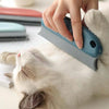 A hand is using a gray pet hair remover brush on the fur of a white cat.
