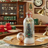 A glass message bottle decorated with a Christmas tree and glitter sits on a gold plate in a festive setting.