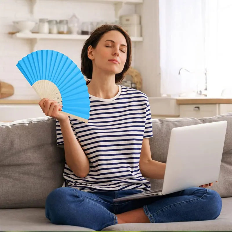 Une femme tient un éventail bleu pliable en bambou et papier, assise sur un canapé.
