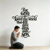 A man is kneeling in front of a large black metal cross wall art featuring the verse For God so loved the world that he gave 