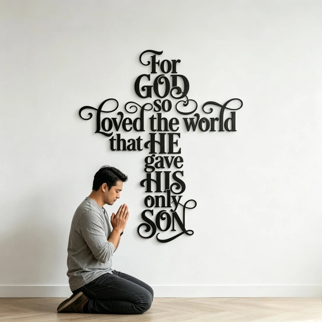A man is kneeling in front of a large black metal cross wall art featuring the verse For God so loved the world that he gave 