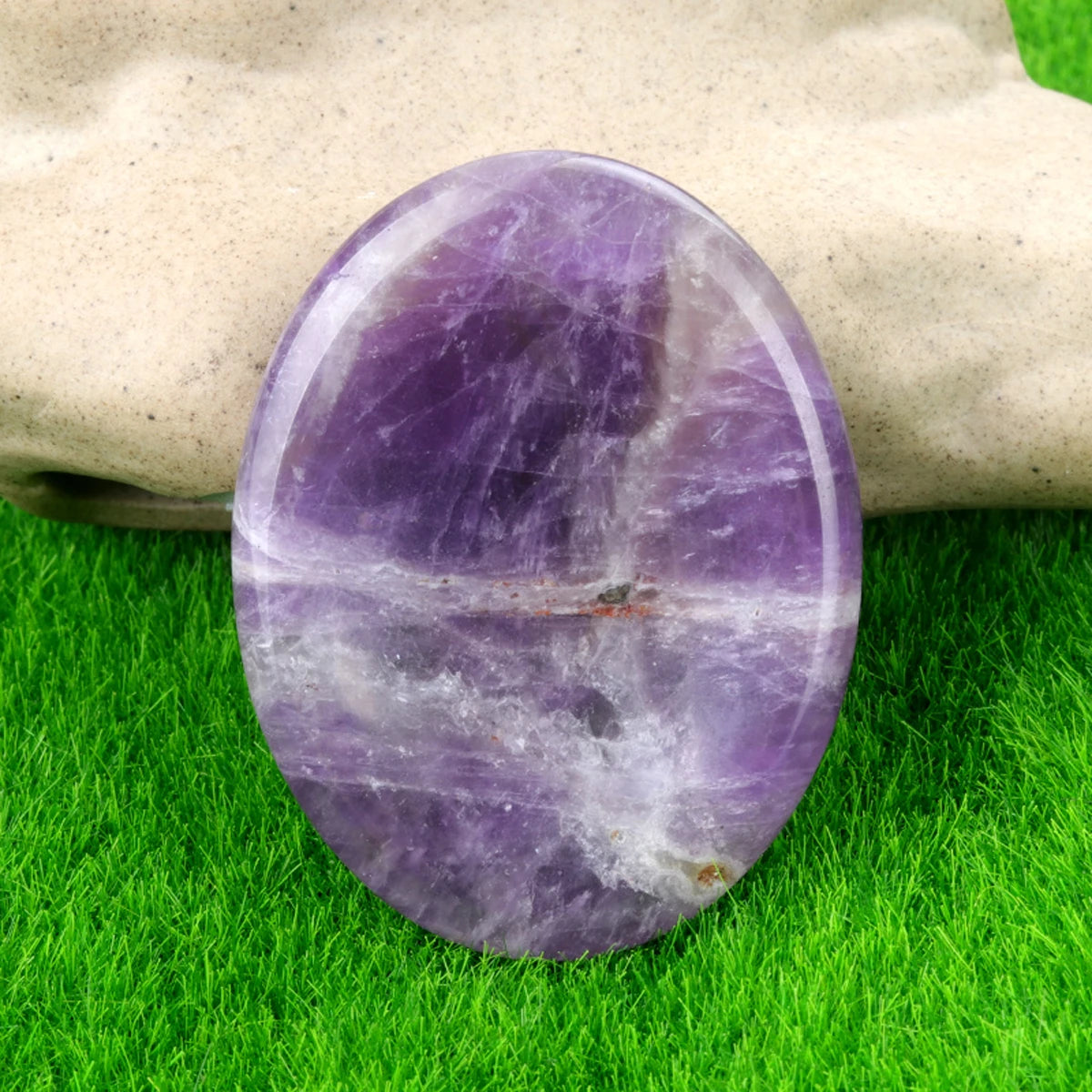 A polished purple natural quartz thumb stone resting on green grass and a hand.