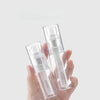A hand holds two clear refillable styling mist spray bottles against a light gray background.