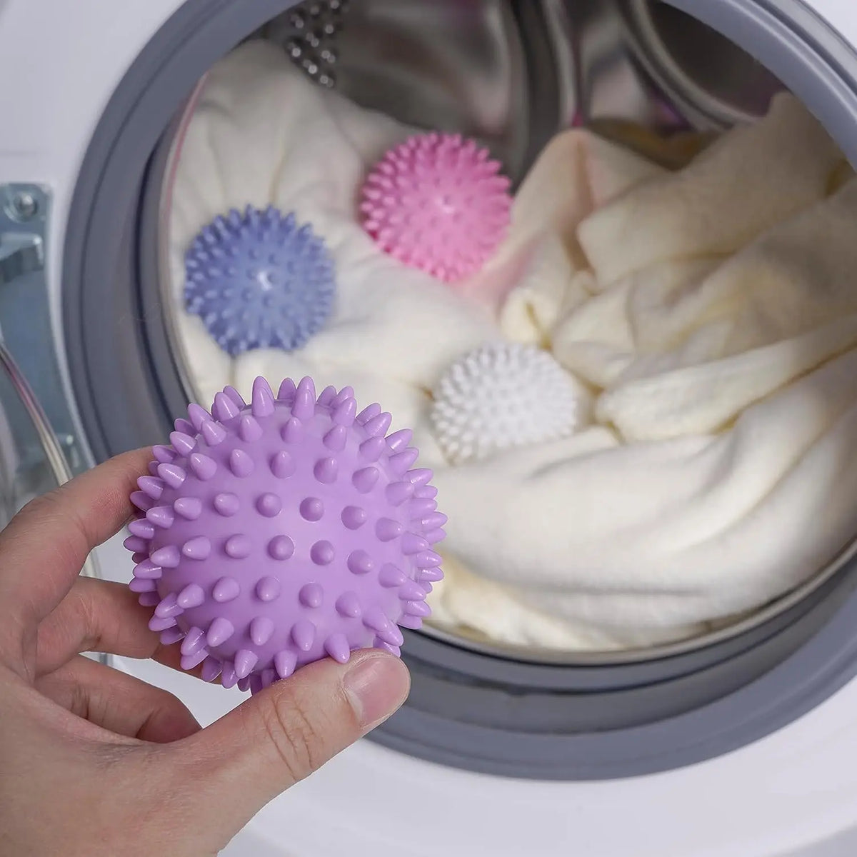 A hand holds a purple laundry ball next to other colored balls inside a laundry basket.