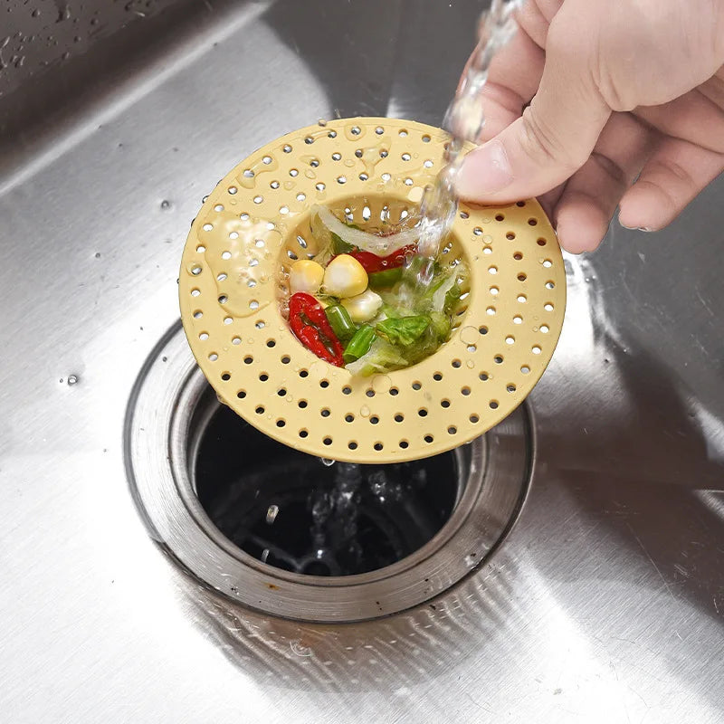 A hand is pouring water through a yellow kitchen sink drain strainer and hair catcher into a metal sink.