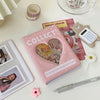 Álbum de fotos en forma de corazón rosa titulado 'Collect' con varios objetos pequeños y un lápiz de color claro cercano.