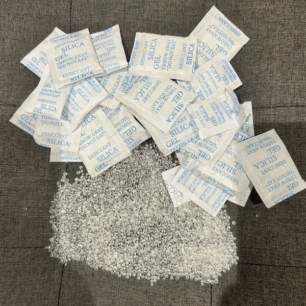 A pile of small white silica gel moisture absorber bags is spread on a dark surface.
