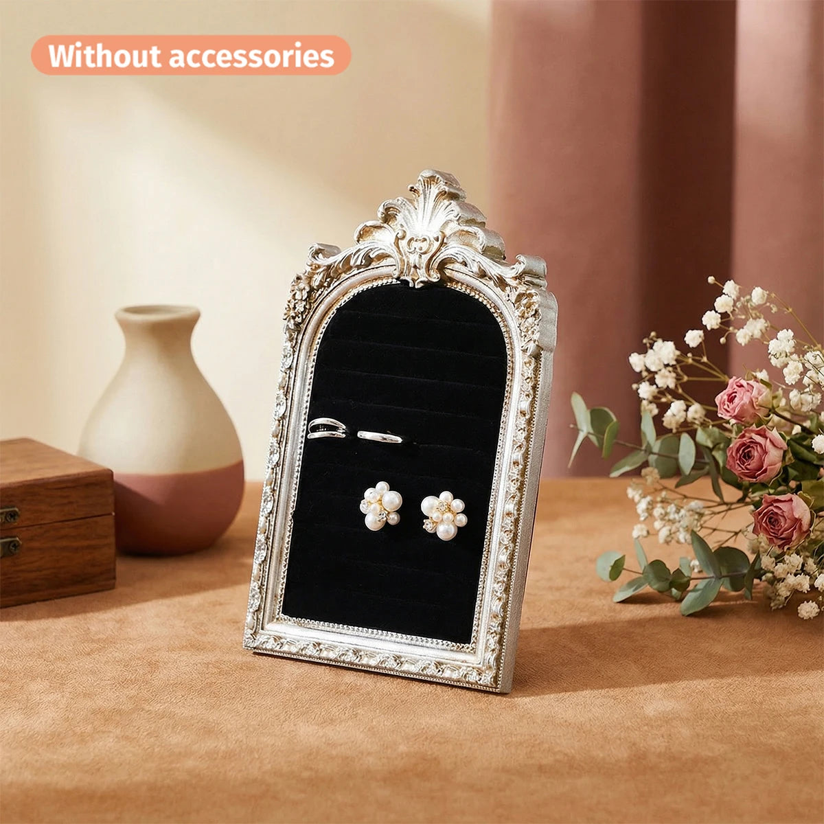 Présentoir de bijoux à cadre argenté avec miroir incurvé sur une surface marron à côté d'un vase et de fleurs.