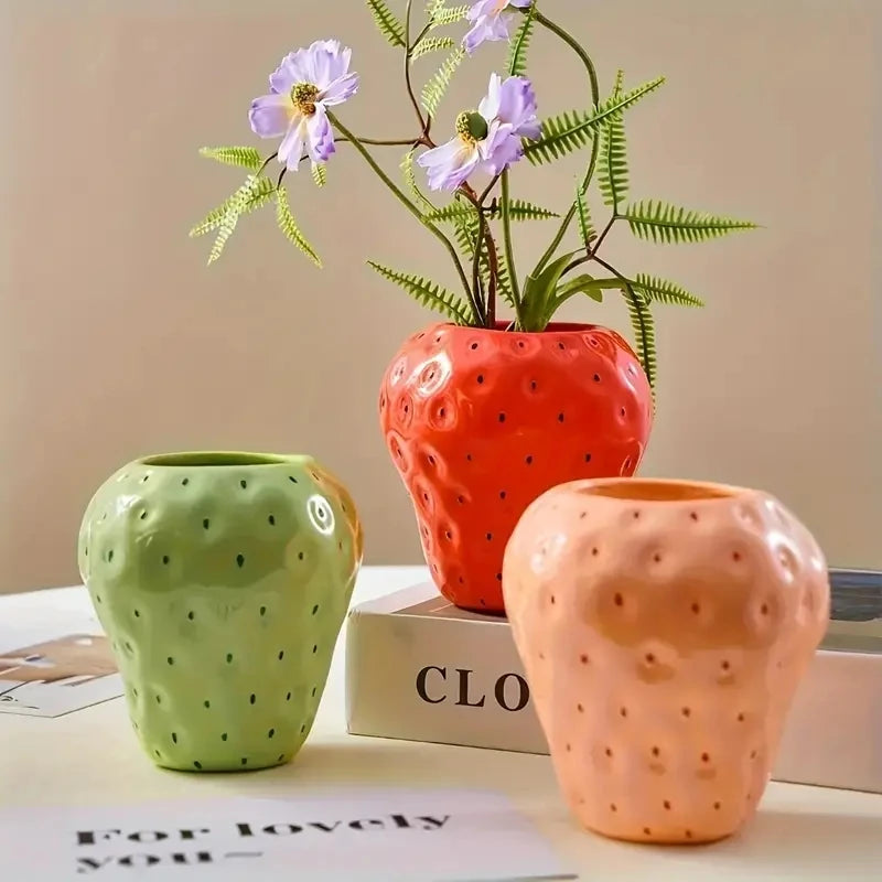 Terrakottafarbenes Harzblumenvase in Form einer Erdbeere, die violette Blumen hält, auf einer weißen Oberfläche präsentiert.