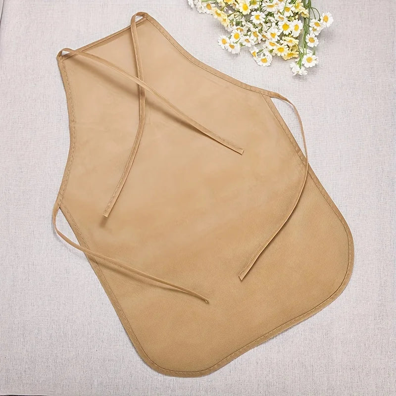 A tan non-woven craft apron with thin straps is displayed on a light fabric next to some white and yellow flowers.