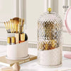 A clear spherical makeup brush organizer with a gold lid sits on a white tray next to various makeup brushes.