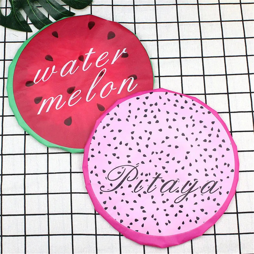 Two reusable fruit shower caps, one red with water melon text and one pink with Pitaya text, are displayed on a black grid ba