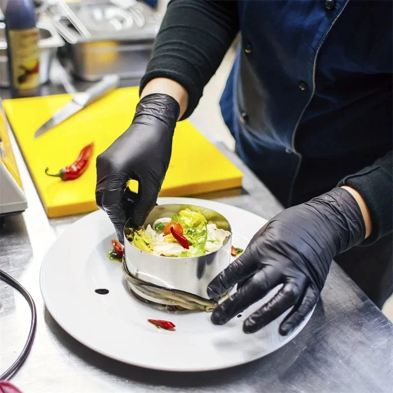 A person wearing black nitrile gloves is carefully placing a piece of food onto a plate in a kitchen setting.