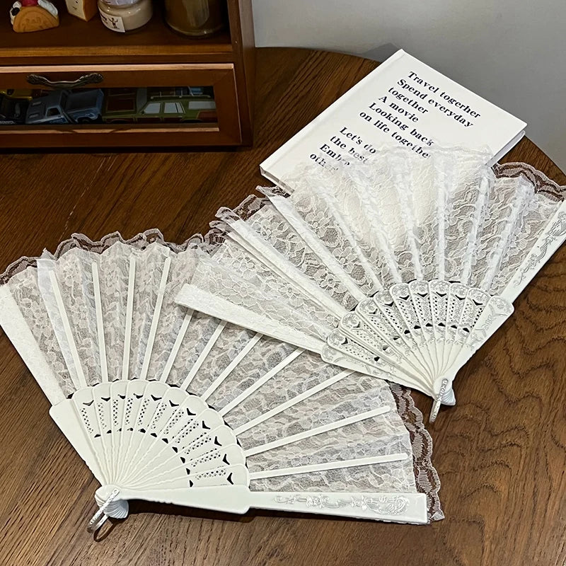 A white lace fabric folding hand fan with a brown edge is displayed on a wooden surface.