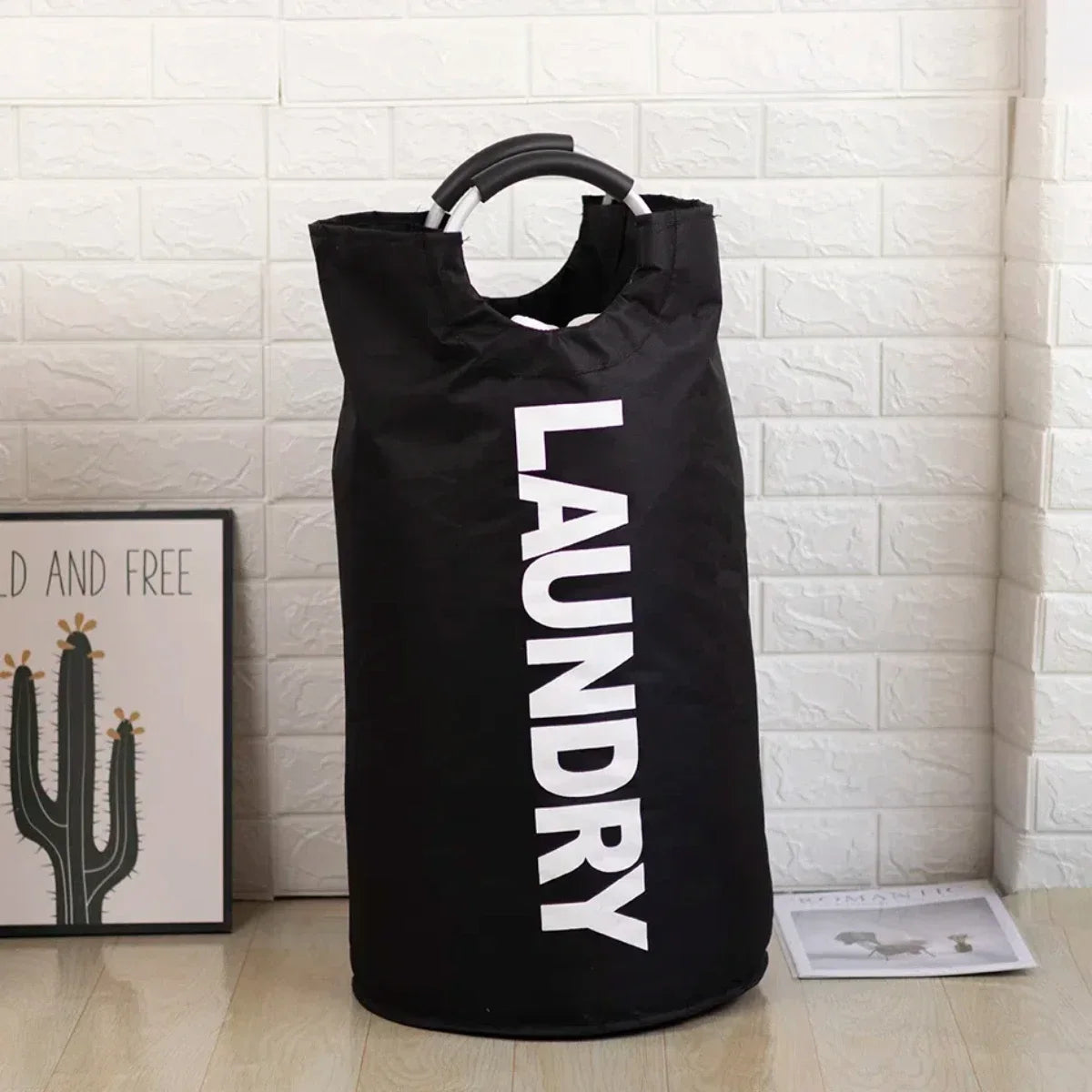 A black foldable laundry basket with a padded handle is displayed against a white brick wall.