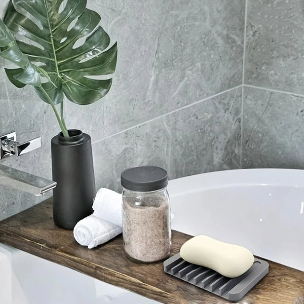 A glass jar of exfoliating scrub and a white silicone soap dish are placed on a wooden vanity next to a sink.