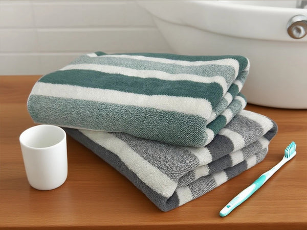 A stack of green and white striped quick-dry bath towels rests on a wooden surface next to a white cup and a toothbrush.