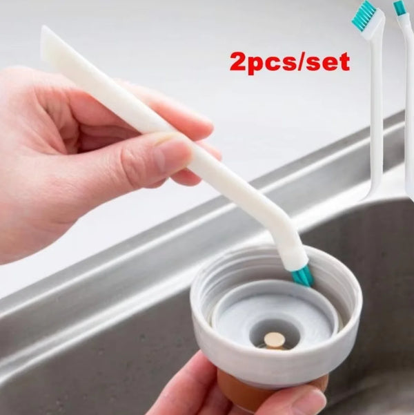 A hand is using a white dual-sided silicone cleaning brush set to clean a bottle inside a stainless steel sink.