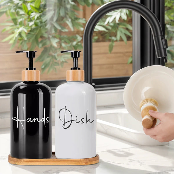 Two refillable hand soap dispenser bottles, one black and one white, sit on a wooden tray next to a kitchen sink.