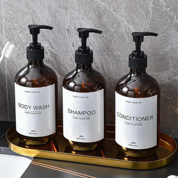 Three dark amber glass bottles of body wash shampoo and conditioner with black pump dispensers displayed on a gold tray