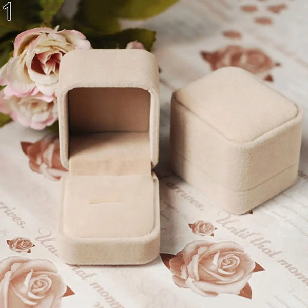 A light beige velvet jewelry box is shown open, with a closed version next to it on a floral patterned surface.