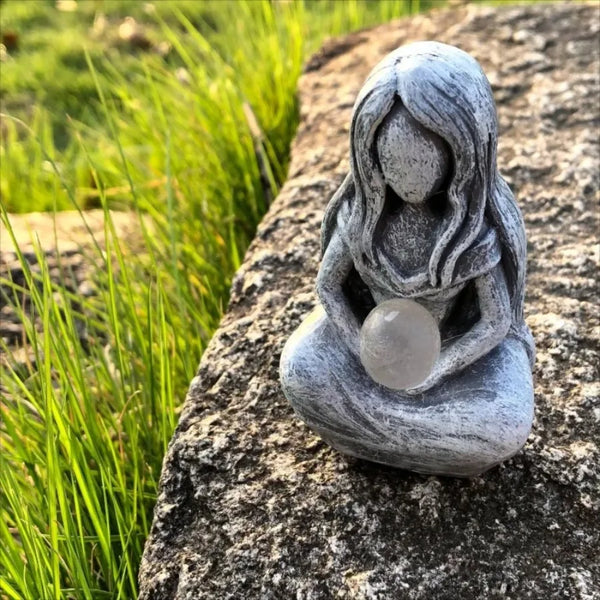 A smooth, grey resin statue of a woman with long hair in a meditative pose rests on a large rock amidst green grass.