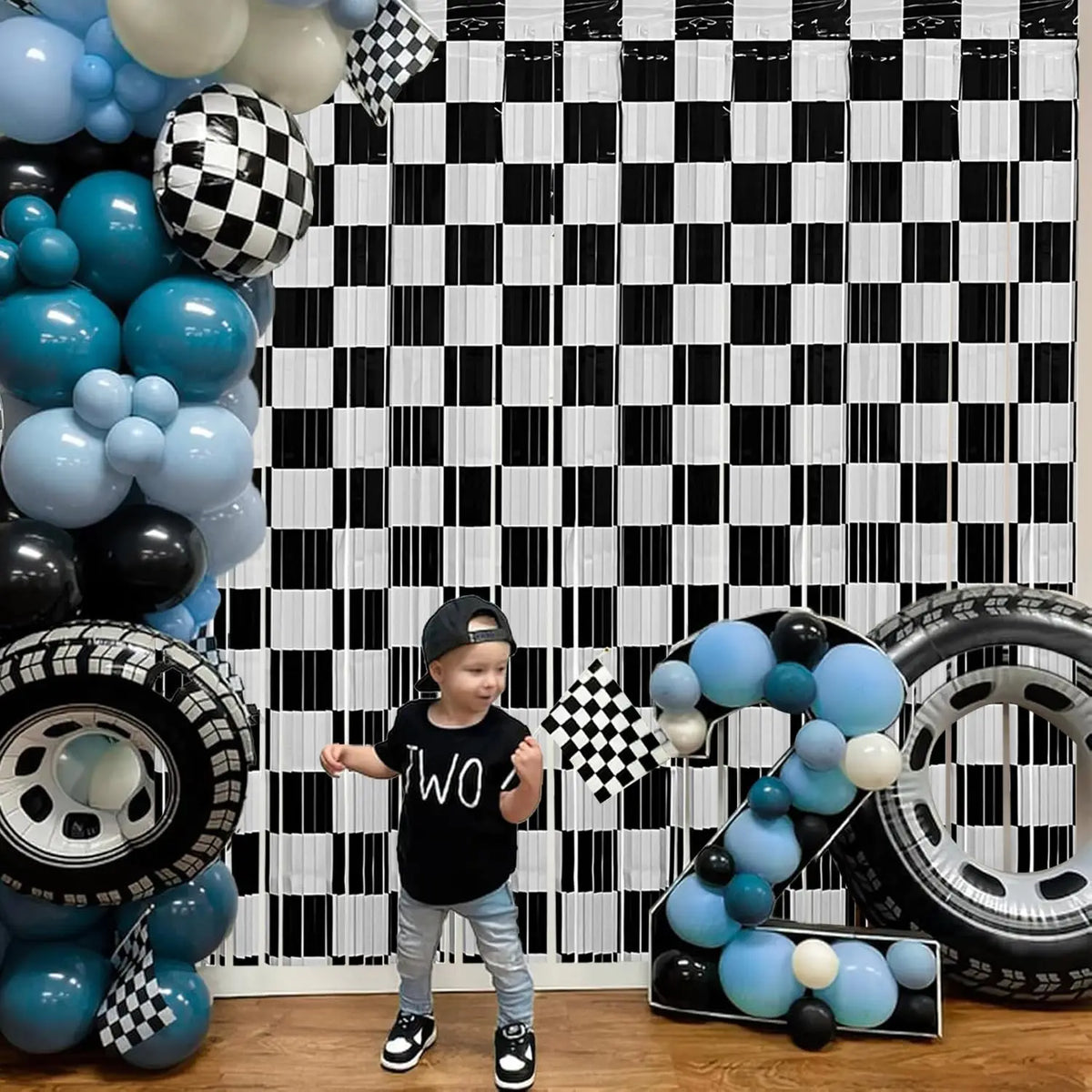 A young boy stands in front of a black and white checkered foil curtain backdrop decorated with blue and black balloons and t