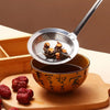 A stainless steel fat skimmer strainer is shown above a bowl of dark liquid and several dried red chili peppers on a wooden b