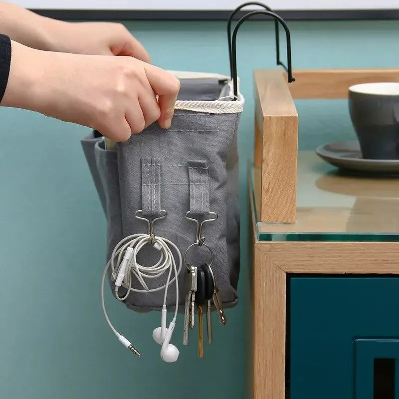 A hand is hanging a grey fabric storage basket with various wires and tools from a black hook on a teal wall.
