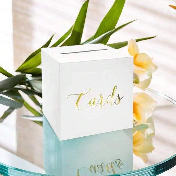 A white elegant wedding card box set with gold lettering sits on a glass surface surrounded by green foliage and yellow flowe