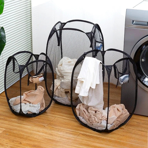 Black collapsible mesh laundry basket with side pockets filled with white and beige laundry on a wooden floor.