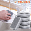 A hand is using a gray double-sided cleaning sponge wipe on a white plate in a sink, with other wipes stacked nearby.