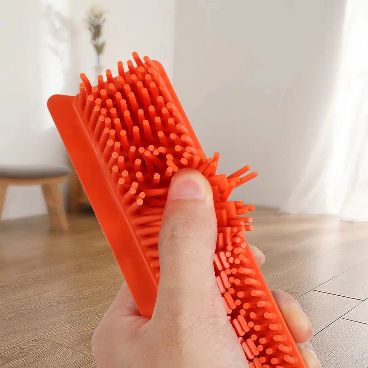 A hand holds an orange adjustable rubber broom with squeegee bristles for pet hair and carpet cleaning.