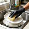 A hand wearing a black nitrile disposable glove is placing a piece of bread on a white plate in a stainless steel sink.