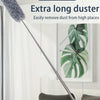 A long, retractable duster brush with a fluffy head is shown next to a potted plant in a bright room.