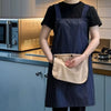 A person wearing a dark blue striped kitchen apron over a black shirt, holding a beige hand towel.