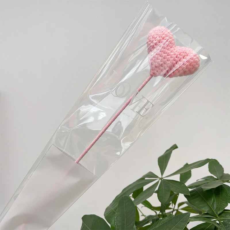 Une fleur en forme de cœur tricotée rose est présentée dans un emballage plastique transparent.