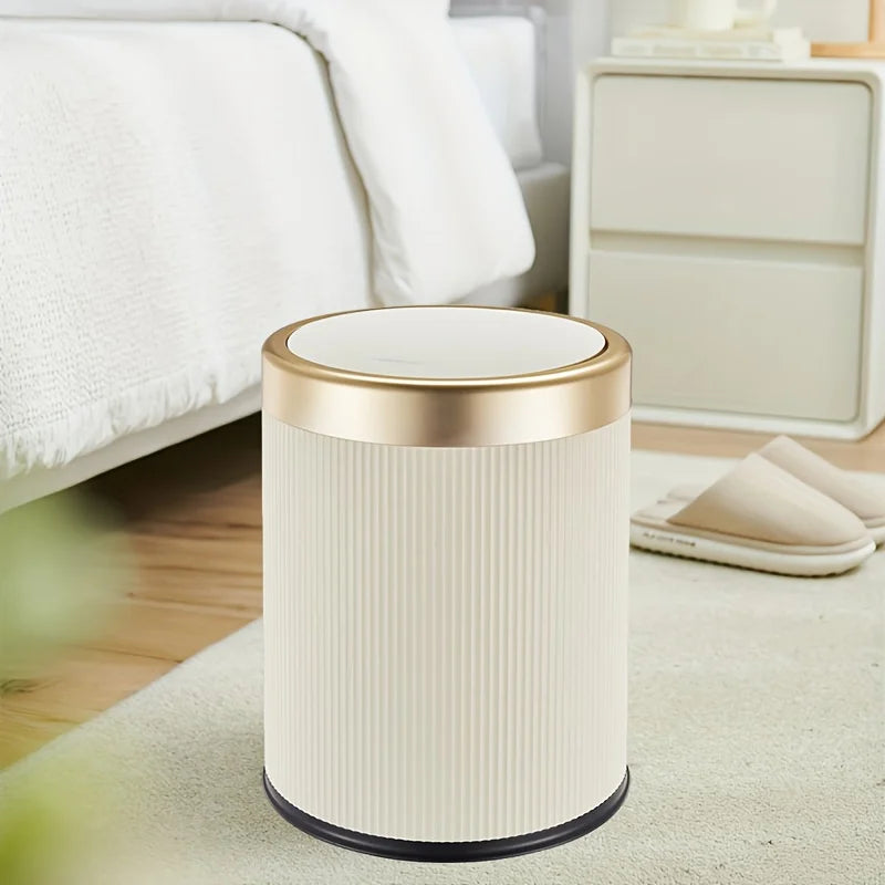 A beige durable press-type trash can with a gold rim sits on a light surface next to a bed.