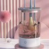 A clear glass container holds a pink liquid with makeup brushes submerged, topped by a white base and three blue cleaning too