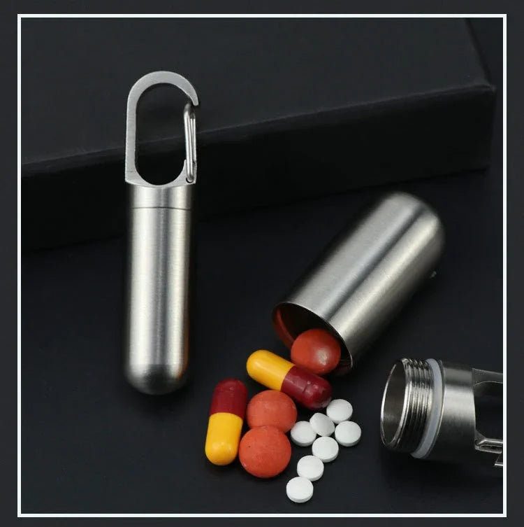 A stainless steel travel pill box and first aid container with a hinged lid and several pills on a dark surface.