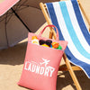 A pink laundry bag rests on a wooden folding chair next to a striped towel and an open umbrella on a sandy surface.