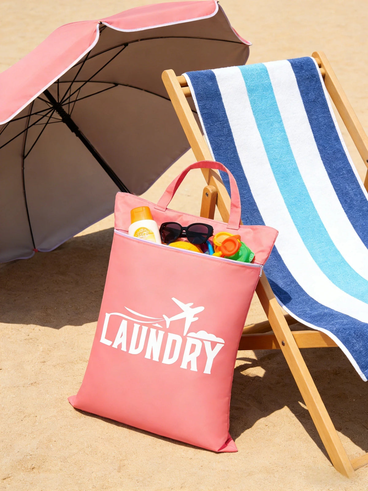 A pink laundry bag rests on a wooden folding chair next to a striped towel and an open umbrella on a sandy surface.