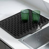 A black silicone drain mat is placed on a kitchen sink, holding two green drinking glasses.