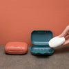 A hand opens a teal and white portable travel soap box against a terracotta-colored wall.